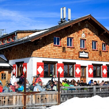 Winterlich verschneite Berghütte mit Holzfassade und zahlreiche Menschen genießen bei Sonnenschein regionale Speisen und Getränke auf der Terrasse | © Archiv Hochwurzenhütte