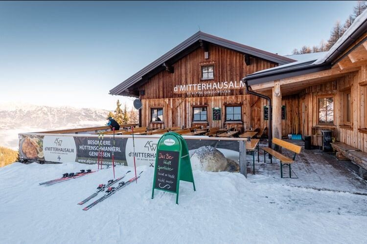 Holzhütte im Schnee mit Terrasse, Skiern davor und Bergblick im Hintergrund. | © Mitterhausalm | Alpine Genusswelten