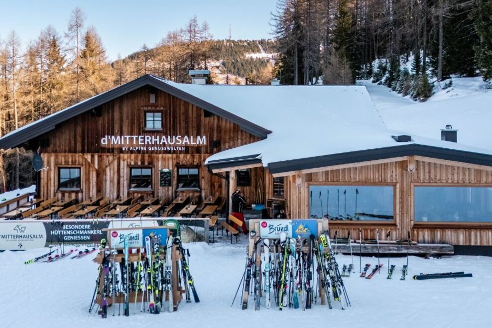 Holzhütte im Schnee mit Terrasse und vielen abgestellten Skiern davor, umgeben von Bäumen. | © Mitterhausalm | Alpine Genusswelten