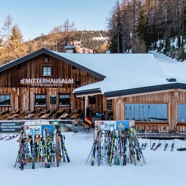 Holzhütte im Schnee mit Terrasse und vielen abgestellten Skiern davor, umgeben von Bäumen. | © Mitterhausalm | Alpine Genusswelten