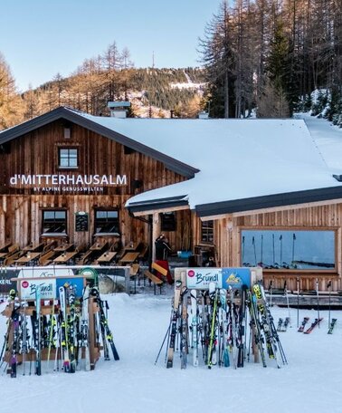 Holzhütte im Schnee mit Terrasse und vielen abgestellten Skiern davor, umgeben von Bäumen. | © Mitterhausalm | Alpine Genusswelten