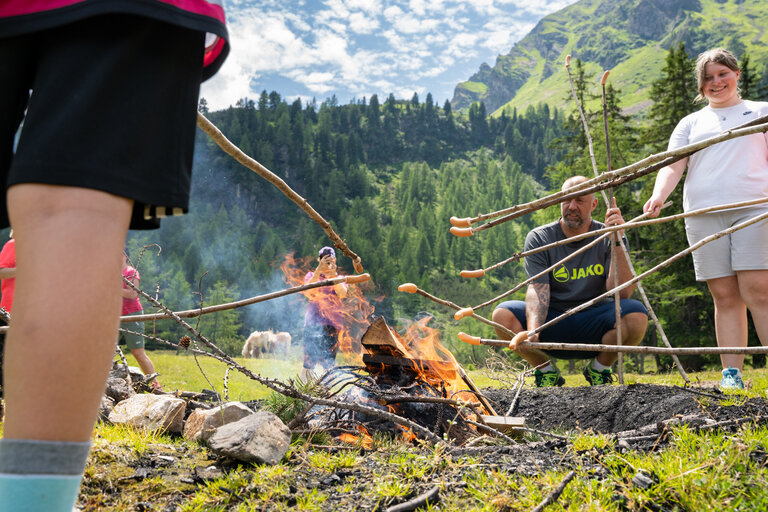 Gruppe grillt Würstchen am Lagerfeuer auf einer Almwiese in den Bergen. | © TVB/Laura Passenbrunner