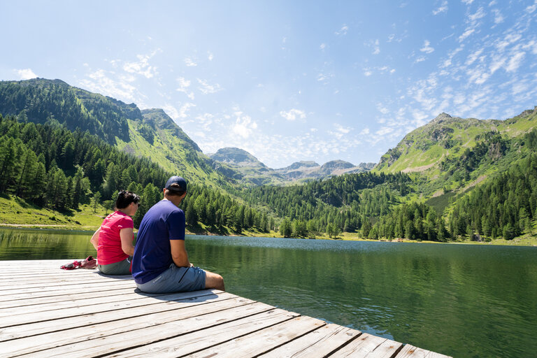 Zwei Personen sitzen am Steg und blicken über einen ruhigen Bergsee inmitten grüner Berge. | © TVB/Laura Passenbrunner