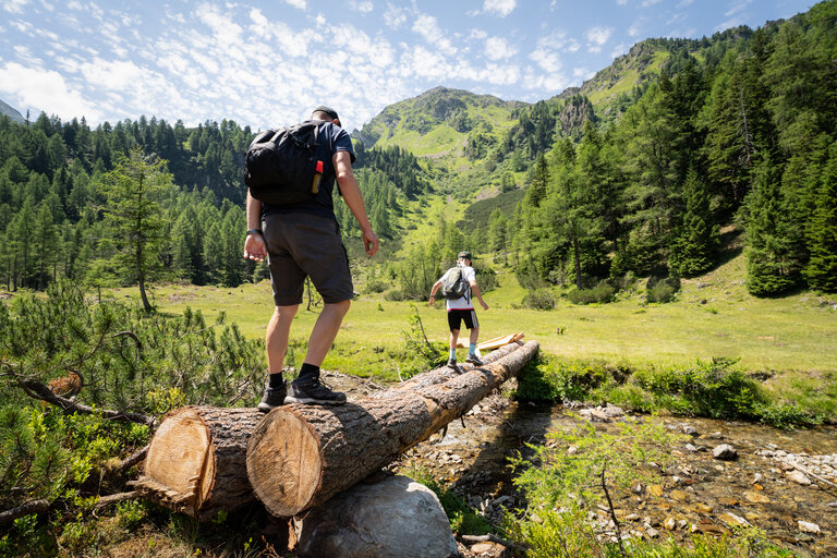 Zwei Wanderer überqueren auf einem Baumstamm einen Bach inmitten alpiner Berglandschaft. | © TVB/Laura Passenbrunner