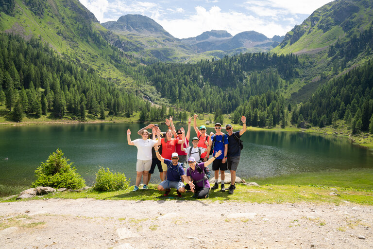 Wandergruppe winkt am Ufer eines Bergsees vor grüner Bergkulisse in die Kamera. | © TVB/Laura Passenbrunner