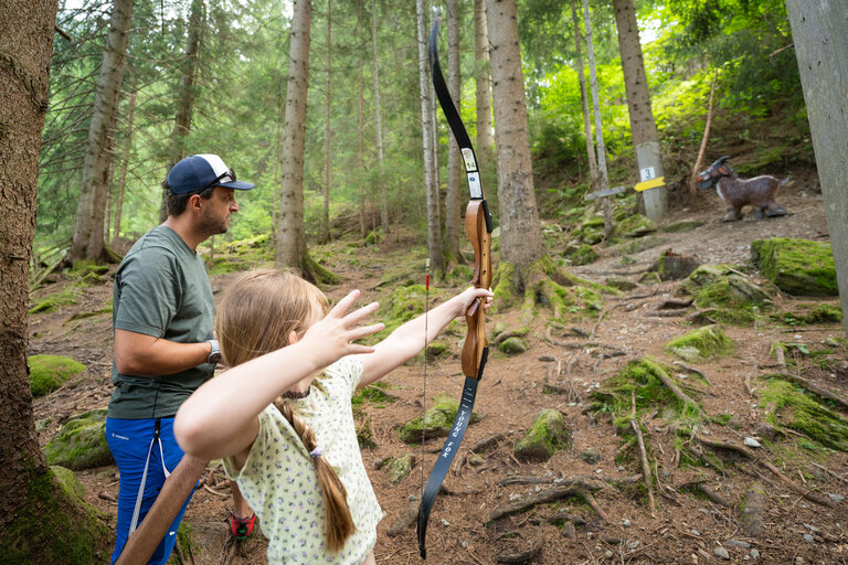 Mädchen zielt mit Bogen auf 3D-Ziel im Wald, betreut von einem Erwachsenen. | © TVB/Laura Passenbrunner