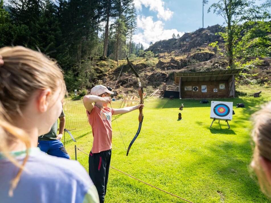 Kind beim Bogenschießen auf einer Wiese, andere Kinder schauen zu. | © TVB/Laura Passenbrunner
