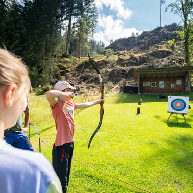 Kind beim Bogenschießen auf einer Wiese, andere Kinder schauen zu. | © TVB/Laura Passenbrunner