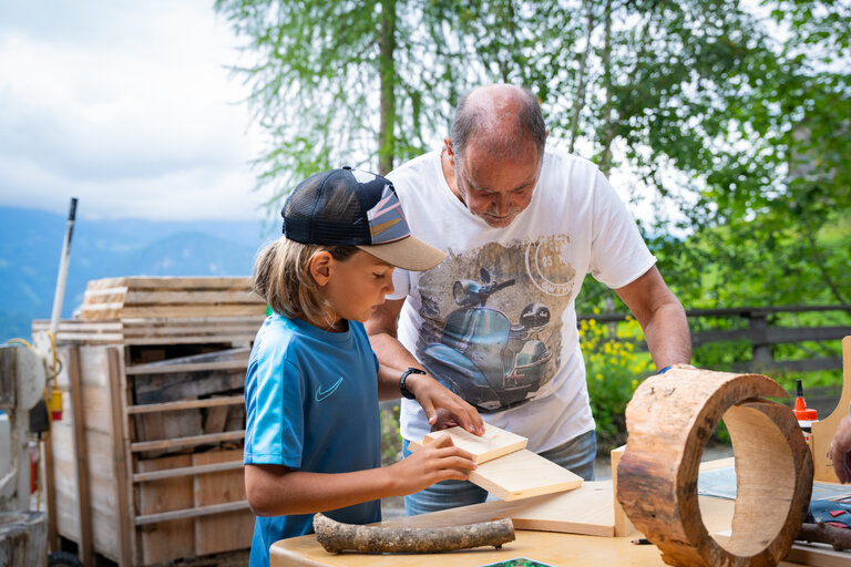Kind und Mann arbeiten gemeinsam an einem Holzstück bei der Kindertischlerei im Freien. | © TVB/Laura Passenbrunner