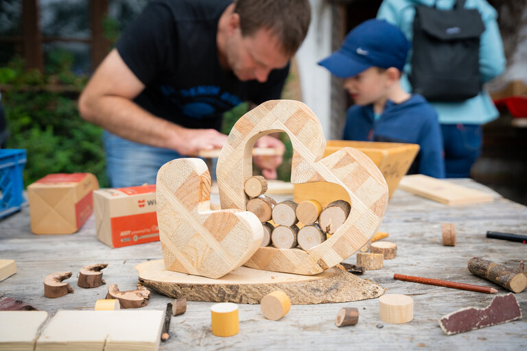 Herzförmige Werkstück auf einer Werkbank, im Hintergrund arbeiten Vater und Kind an einem Bastelprojekt. | © TVB/Laura Passenbrunner
