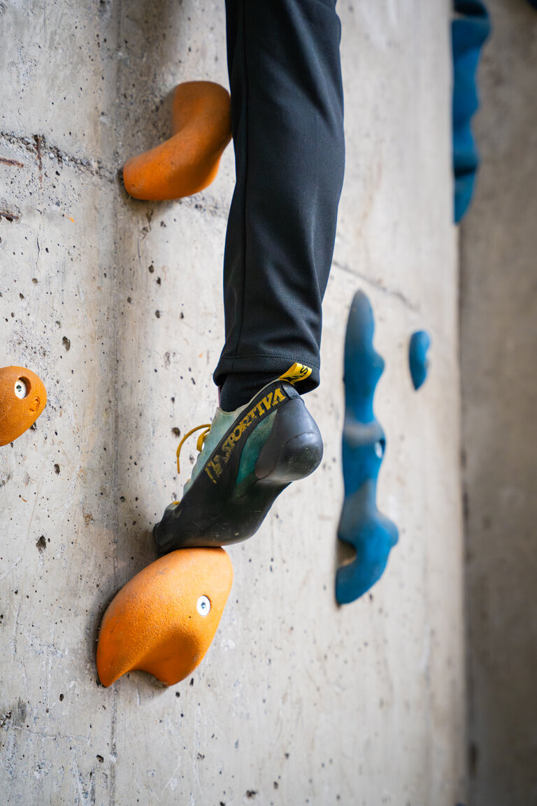 Nahaufnahme eines Kletterschuhs auf orangefarbenem Klettergriff an Betonwand. | © TVB/Laura Passenbrunner