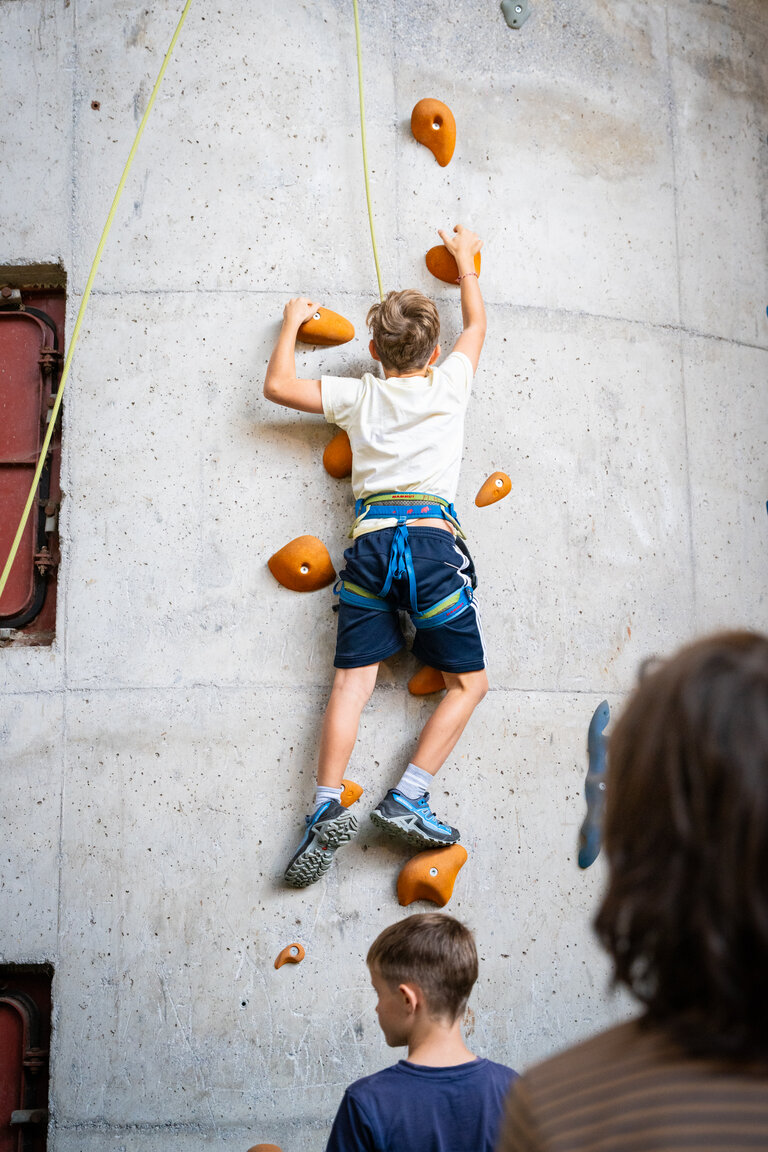 Ein Junge klettert gesichert eine Indoor-Kletterwand mit orangefarbenen Griffen hinauf. | © TVB/Laura Passenbrunner