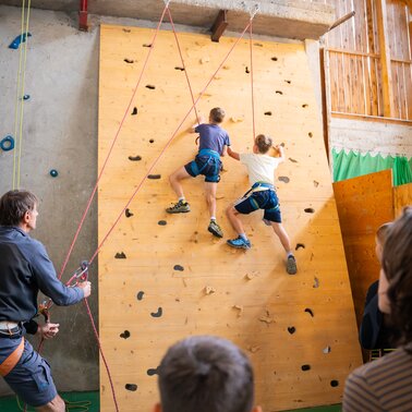 Zwei Kinder klettern gesichert an einer Indoor-Kletterwand, Erwachsene sichern und schauen zu. | © TVB/Laura Passenbrunner