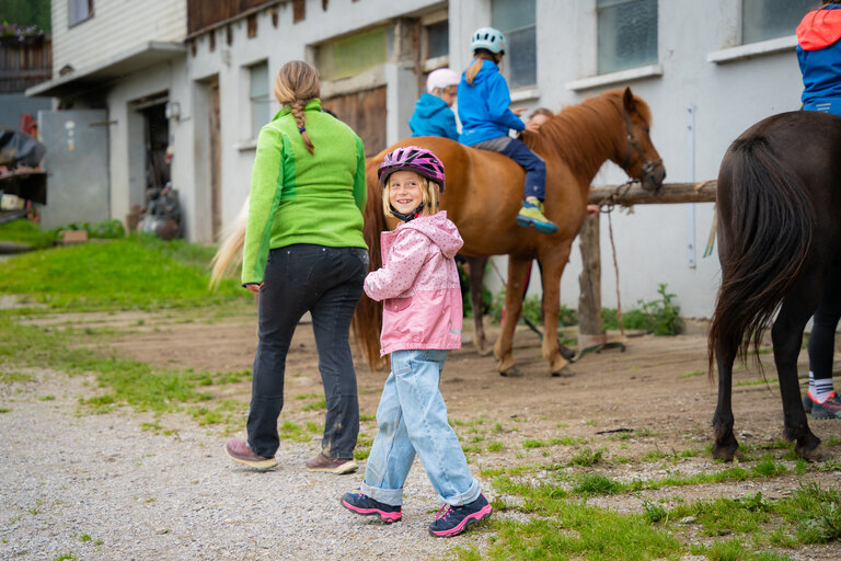 Lächelndes Mädchen am Reitstall, im Hintergrund Kinder auf Pferden. | © TVB/Laura Passenbrunner