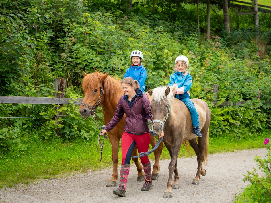 Zwei Kinder reiten geführt durch eine grüne Waldlandschaft. | © TVB/Laura Passenbrunner