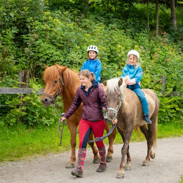 Zwei Kinder reiten geführt durch eine grüne Waldlandschaft. | © TVB/Laura Passenbrunner