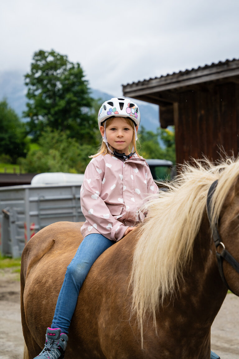 Mädchen mit Helm sitzt auf braunem Pferd vor Stallgebäude. | © TVB/Laura Passenbrunner
