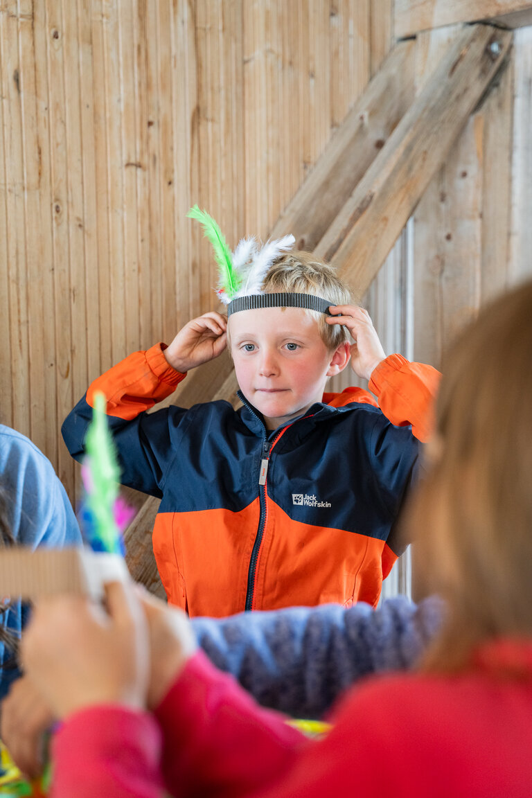 Kind setzt sich ein gebasteltes Stirnband mit bunten Federn auf. | © TVB/Laura Passenbrunner