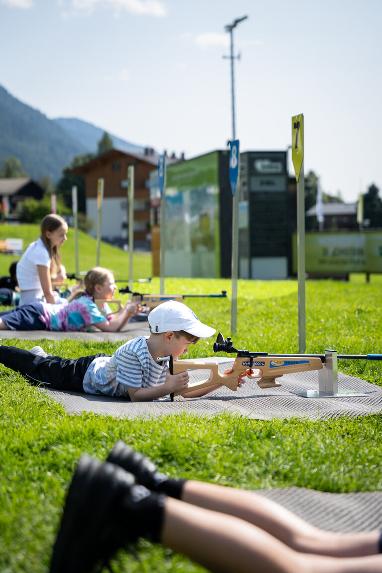 Mehrere Kinder liegen auf Matten auf einer Wiese und zielen mit Lasergewehren auf Zielscheiben beim Kinder-Laserbiathlon. | © TVB/Laura Passenbrunner