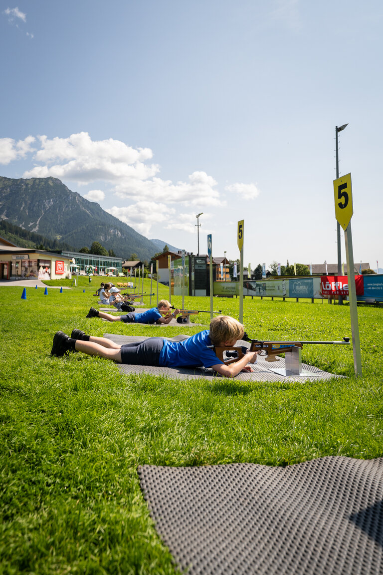 Kinder liegen auf Matten im Gras und üben mit Lasergewehren das Zielen beim Laserbiathlon in Ramsau am Dachstein. | © TVB/Laura Passenbrunner