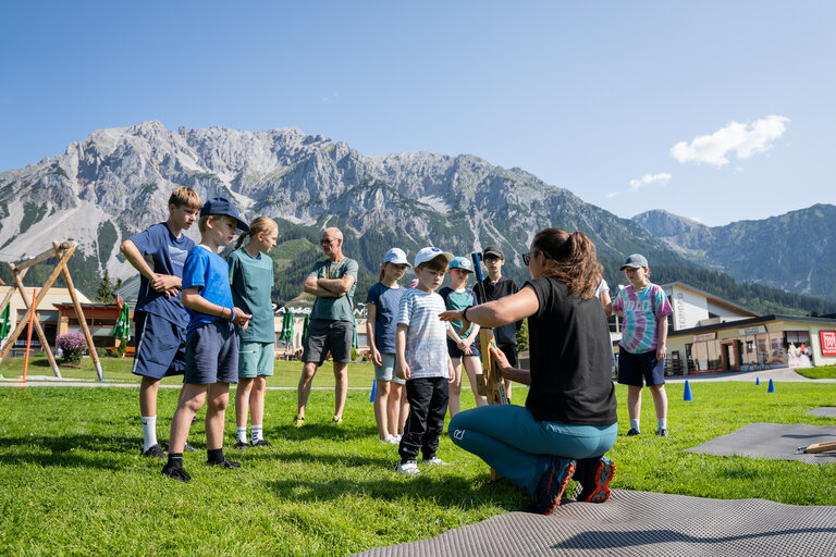Kinder hören auf einer Wiese einer Betreuerin beim Erklären des Laserbiathlons zu, im Hintergrund die Dachstein-Bergkulisse. | © TVB/Laura Passenbrunner