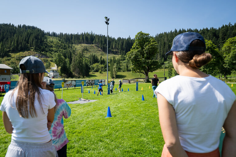 Mehrere Kinder trainieren auf einer Wiese beim Laserbiathlon in Ramsau am Dachstein, während zwei Erwachsene im Vordergrund zusehen. | © TVB/Laura Passenbrunner
