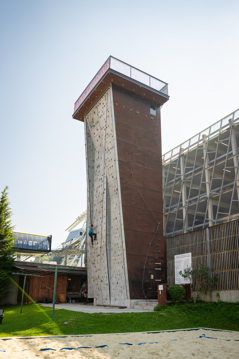 Junge Person klettert gesichert an einer hohen, senkrechten Outdoor-Kletterwand mit bunten Griffen. | © TVB/Laura Passenbrunner