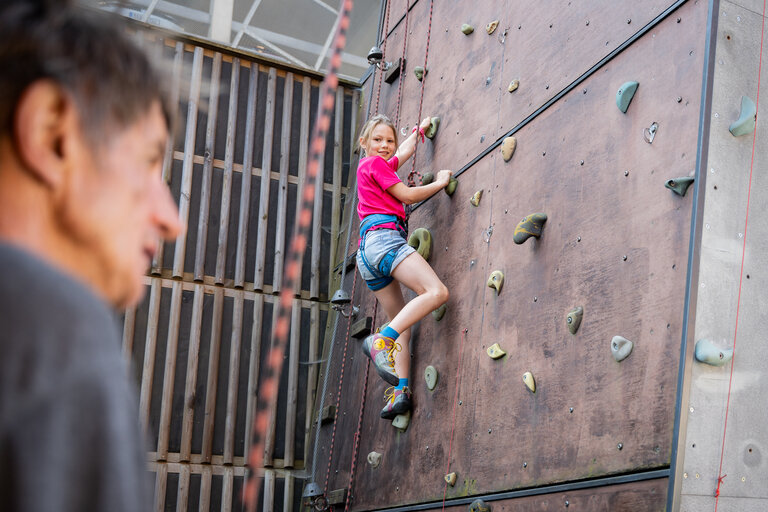 Kind beim gesicherten Klettern an einer hohen künstlichen Kletterwand im Freien. | © TVB/Laura Passenbrunner