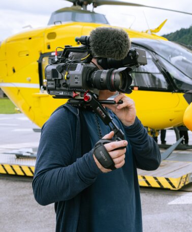 Ein Mann in Bergrettungsoutfit, der von einer Kamera gefilmt wird. Im Hintergrund steht ein Helikopter | © ZDF_2024_17. Staffel