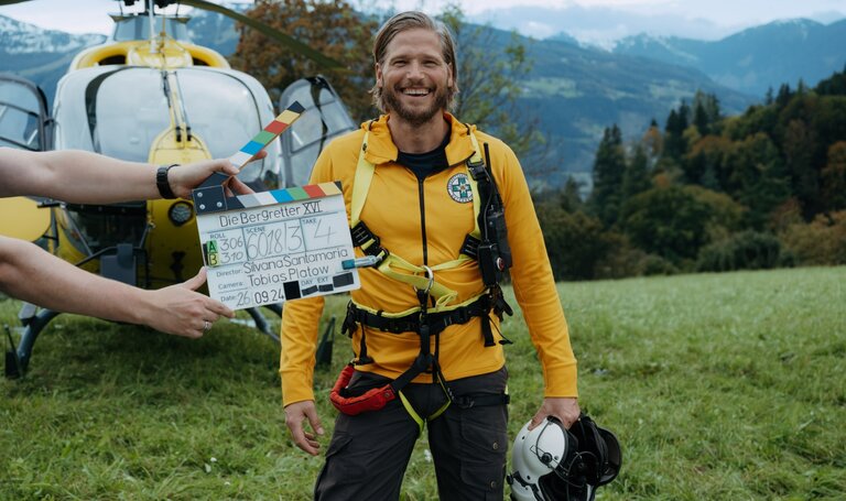 Ein Mann in Bergrettungsoutfit steht vor einem Helikopter, hält eine Filmklappe in der Hand und lacht.  | © ZDF_2024_17. Staffel