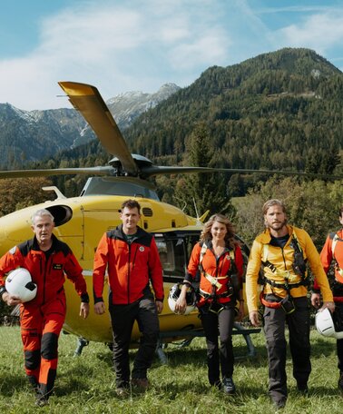 Fünf Personen in Bergrettungsoutfit marschieren auf einer Wiese. Im Hintergrund steht ein Helikopter.  | © ZDF_2024_17. Staffel