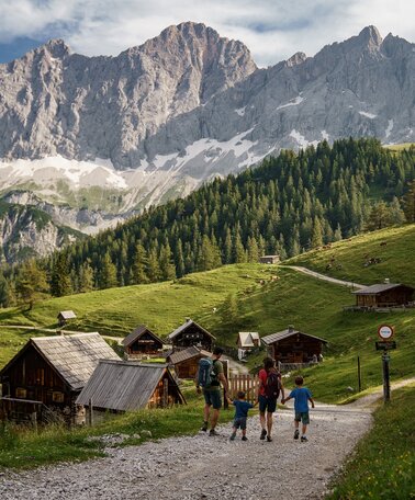 Neustattalm mit dem Dachsteinmassiv im Hintergrund und einer Familie, die in Richtung Neustattalm wandert | © Christine Höflehner