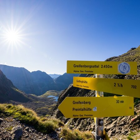 Wegweiser am Greifenbergsattel (2.450 m) mit Blick ins Tal und tiefblauem Bergsee im Gegenlicht der Morgensonne. | © Martin Huber