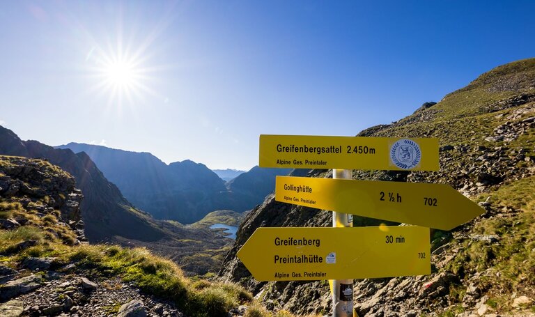 Wegweiser am Greifenbergsattel (2.450 m) mit Blick ins Tal und tiefblauem Bergsee im Gegenlicht der Morgensonne. | © Martin Huber
