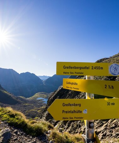 Wegweiser am Greifenbergsattel (2.450 m) mit Blick ins Tal und tiefblauem Bergsee im Gegenlicht der Morgensonne. | © Martin Huber