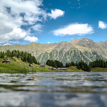 Flache Aufnahme vom Duisitzkarsee mit Almhütten im Hintergrund mit leichten Wolken im Sommer. Das Schilf ragt leicht aus dem See und die Berge im Hintergrund werden von der Sonne angeleuchtet. | © Christine Höflehner