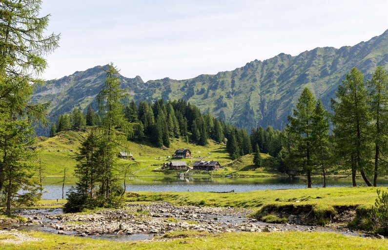 Ein glasklarer Bergsee liegt eingebettet in sanfte Almwiesen, umgeben von urigen Hütten und grünen Berghängen unter blauem Himmel. | © Hans-Peter Steiner