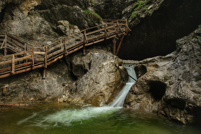 Eine enge Schlucht mit moosbedeckten Felswänden, durch die ein klarer Gebirgsbach rauscht. Holzstege führen direkt durch das felsige Gelände. | © Marlene Eggmayr